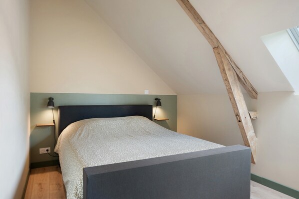 A bedroom with a sloped ceiling, a bed with a patterned bedspread, two bedside lamps, and a small shelf.