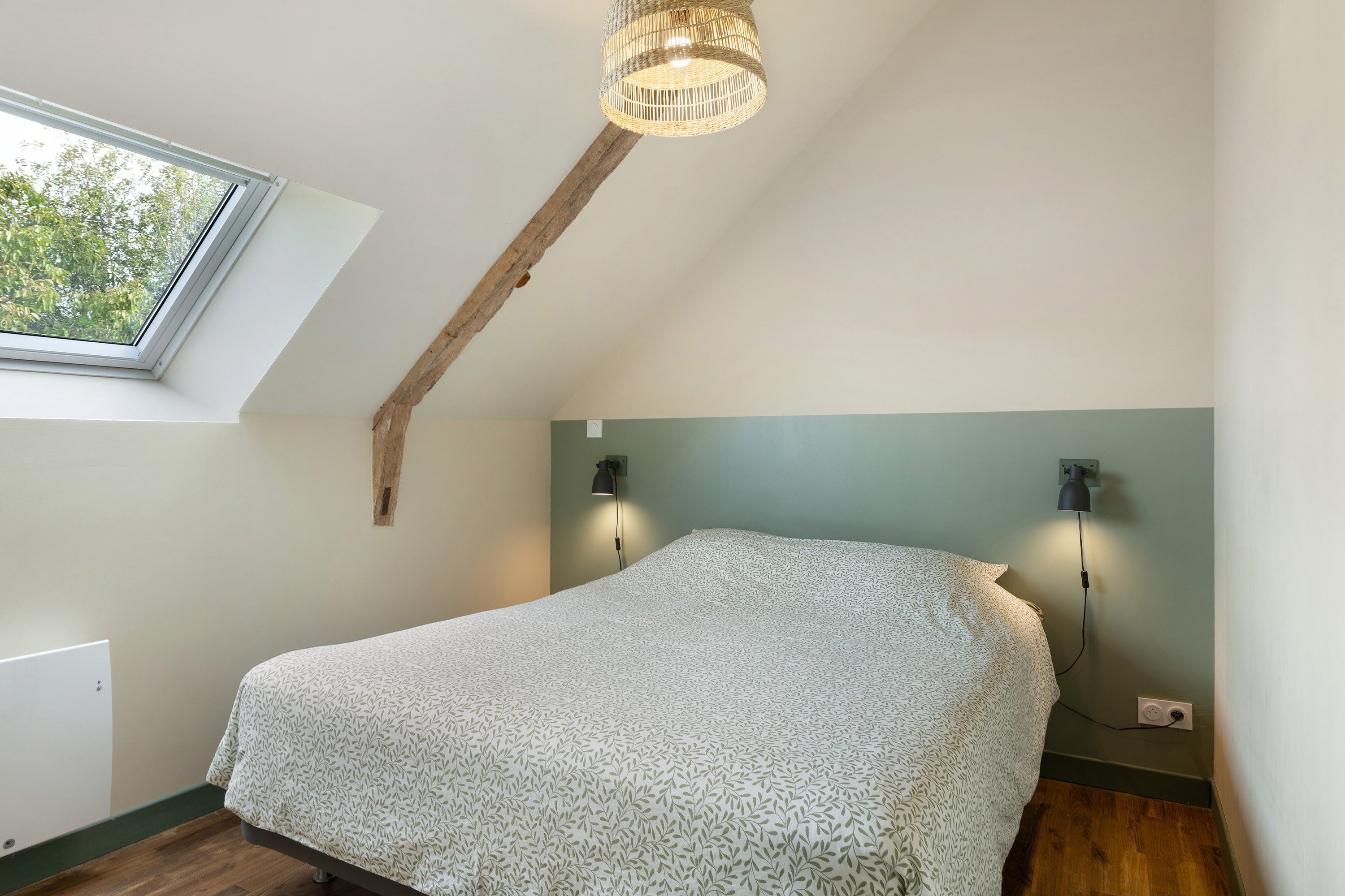 Een slaapkamer met een schuine plafond, een dakraam, een bed met een bedsprei met een patroon, twee wandlampen en een houten balk.