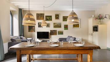A dining area with a wooden table set for four, two hanging pendant lights, a flat-screen TV, and a sofa in the background.