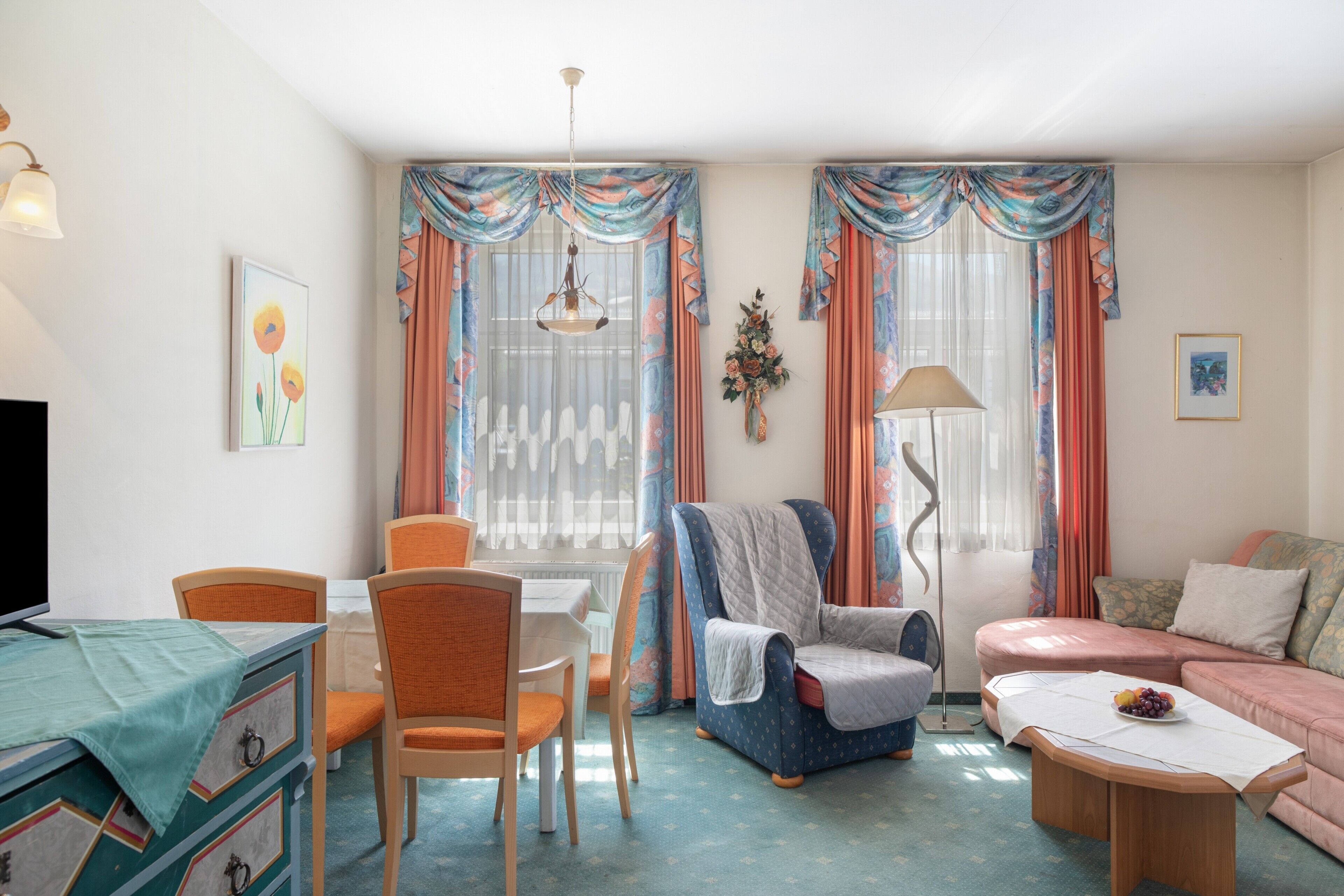 Un salon comprenant un téléviseur, une table à manger avec des chaises, un canapé, un fauteuil, une lampe sur pied et un arrangement floral fixé au mur.