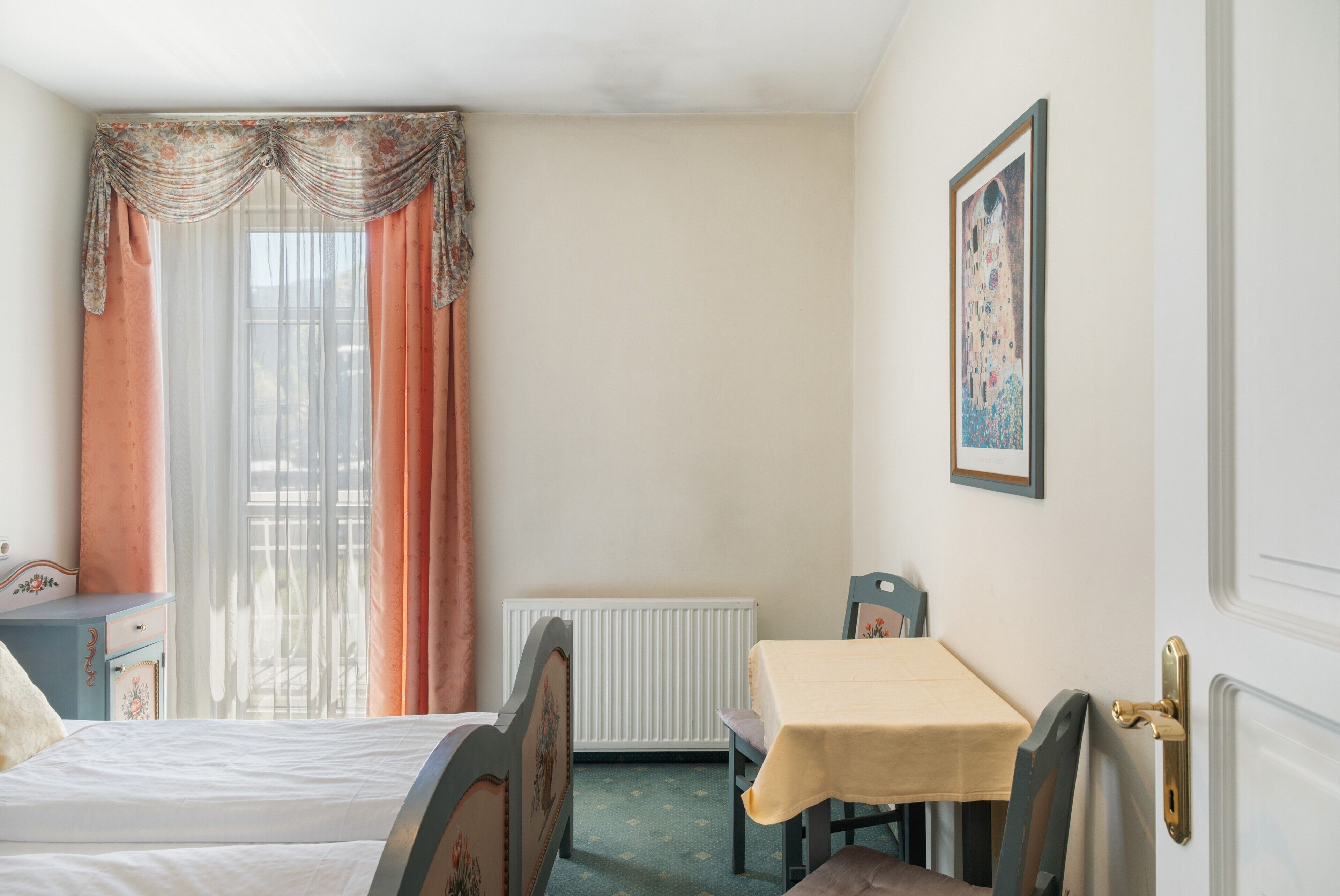 Een slaapkamer met een bed, een tafel, een stoel, een radiator en een schilderij aan de muur.