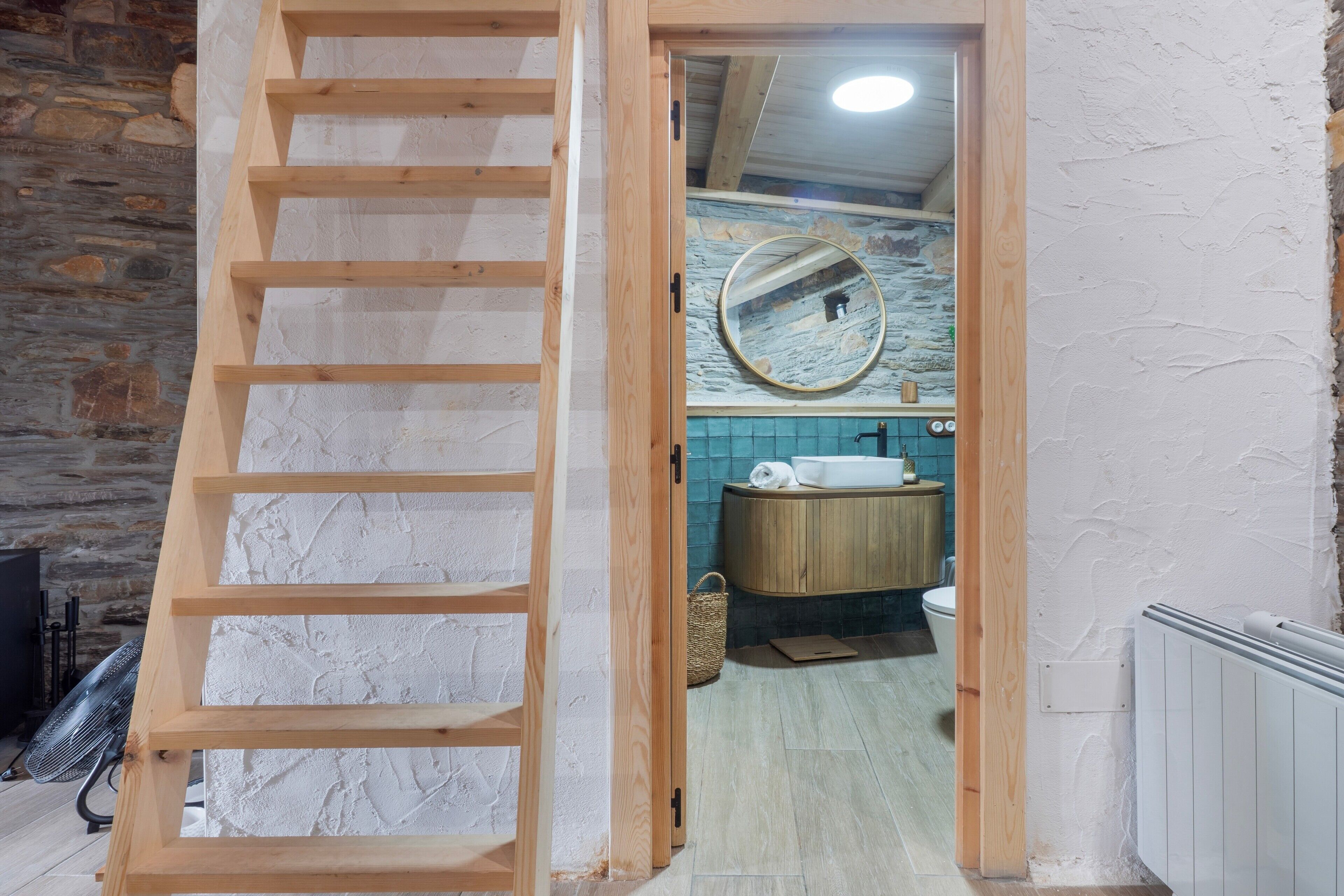 Une échelle en bois mène à une salle de bain avec un mur en pierre, un miroir rond et un meuble-lavabo en bois.