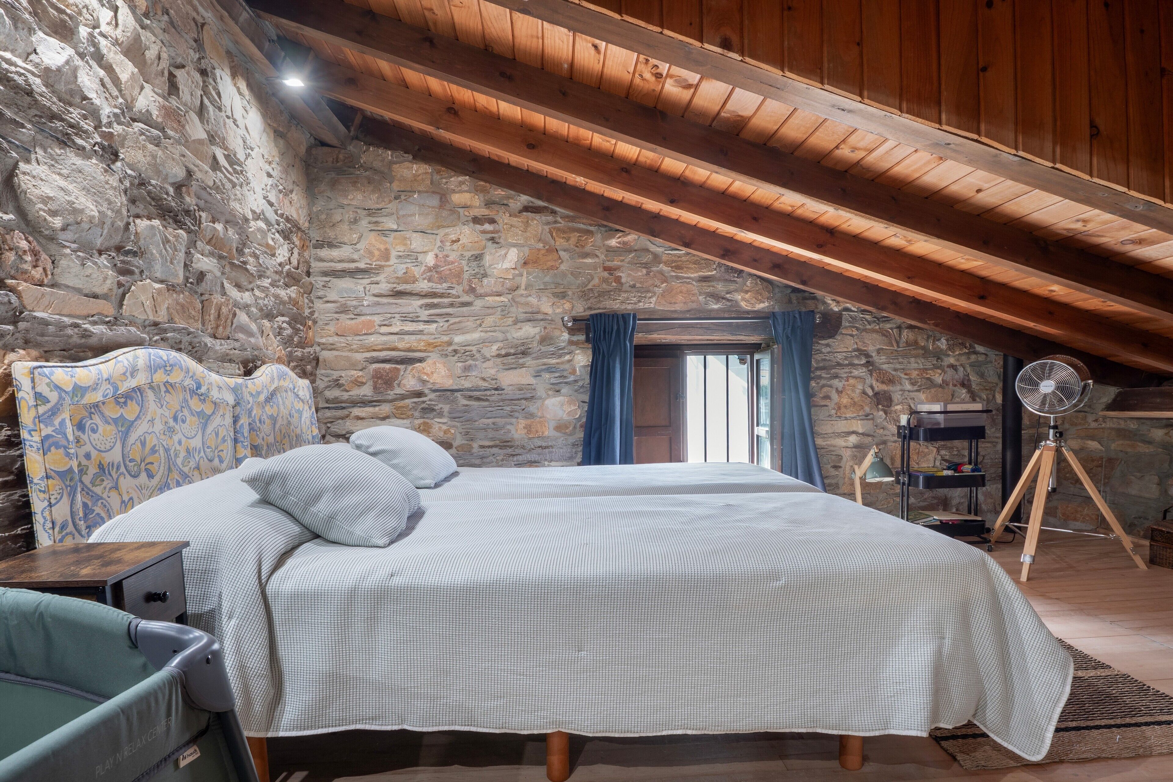 Un dormitorio con pared de piedra, techo de madera, cama con ropa de cama estampada, un escritorio y un ventilador de pie.