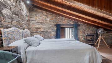 Un dormitorio con pared de piedra, techo de madera, cama con ropa de cama estampada, un escritorio y un ventilador de pie.