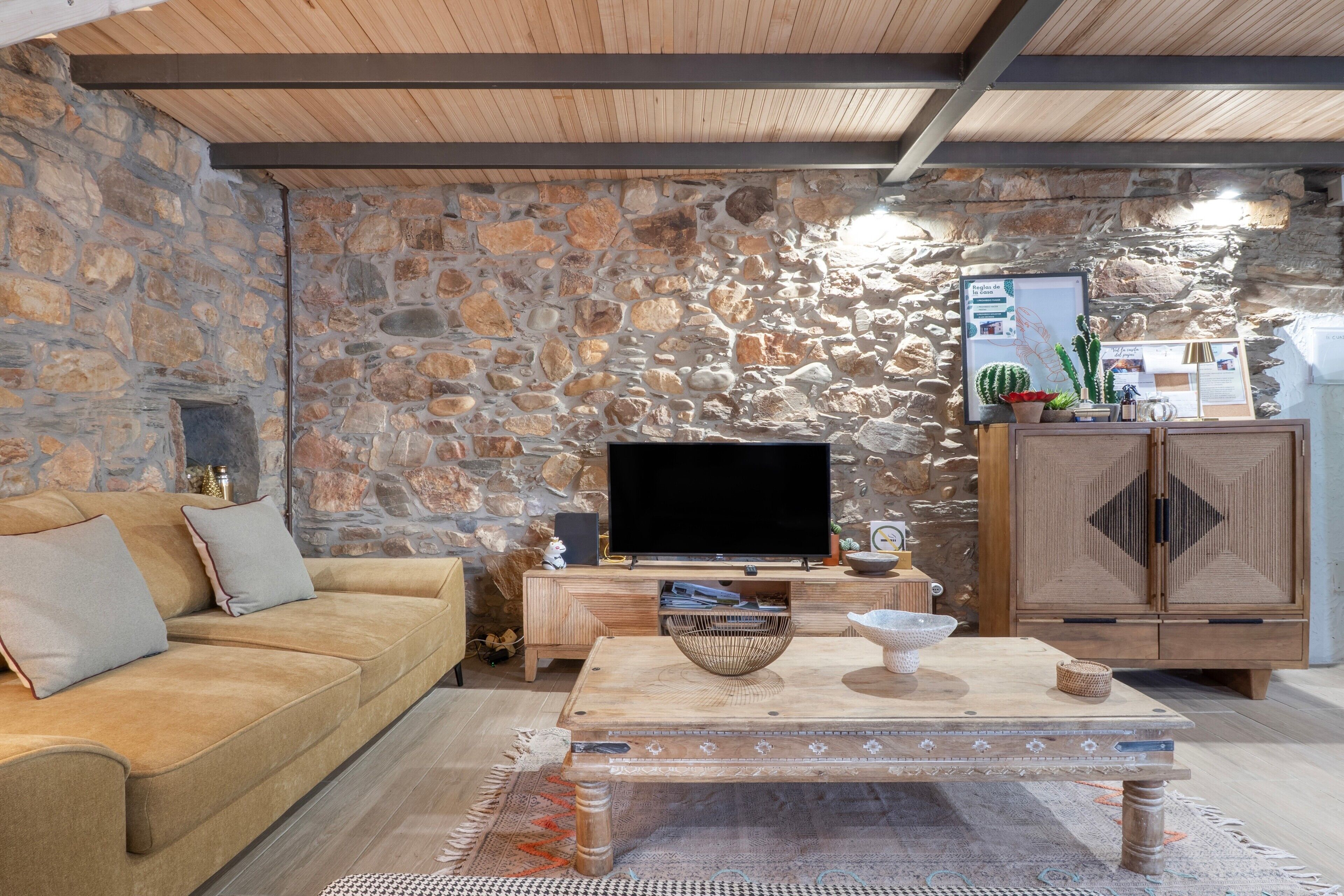 Een woonkamer met een stenen muur, een houten plafond, een bank, een salontafel en een televisie.