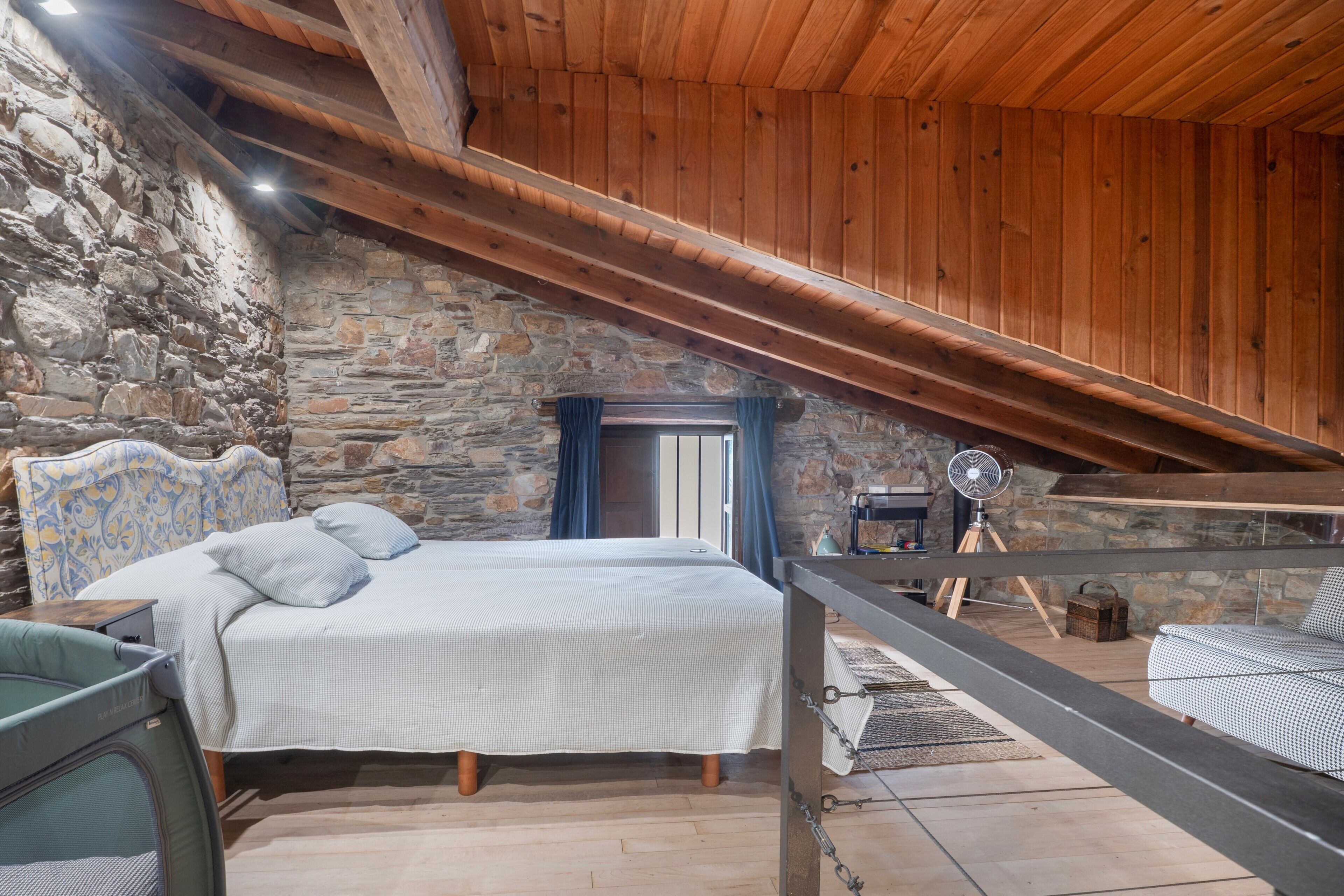 Een slaapkamer met een stenen muur, houten balken in het plafond, een bed met een geborduurde spreidekbed en een ventilator.