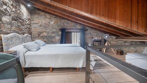 Un dormitorio con pared de piedra, vigas de madera en el techo, una cama con colcha estampada, una mesita pequeña y un ventilador.