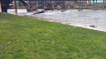 A grassy area with a stone bridge over a river, buildings in the background, and a hill.