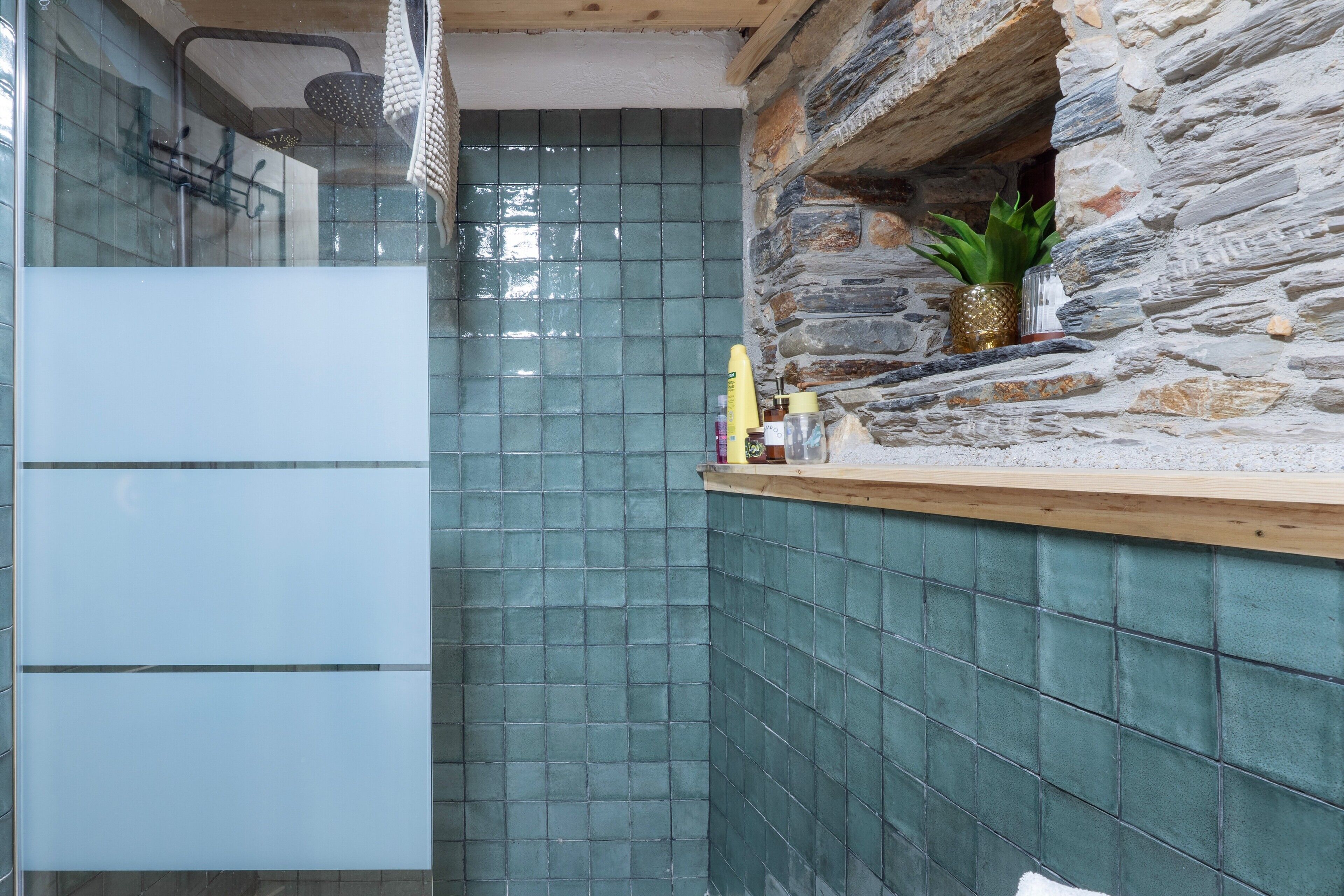Une salle de bain avec une douche, des murs carrelés, une étagère en bois où sont rangés les articles de toilette et un mur en pierre.