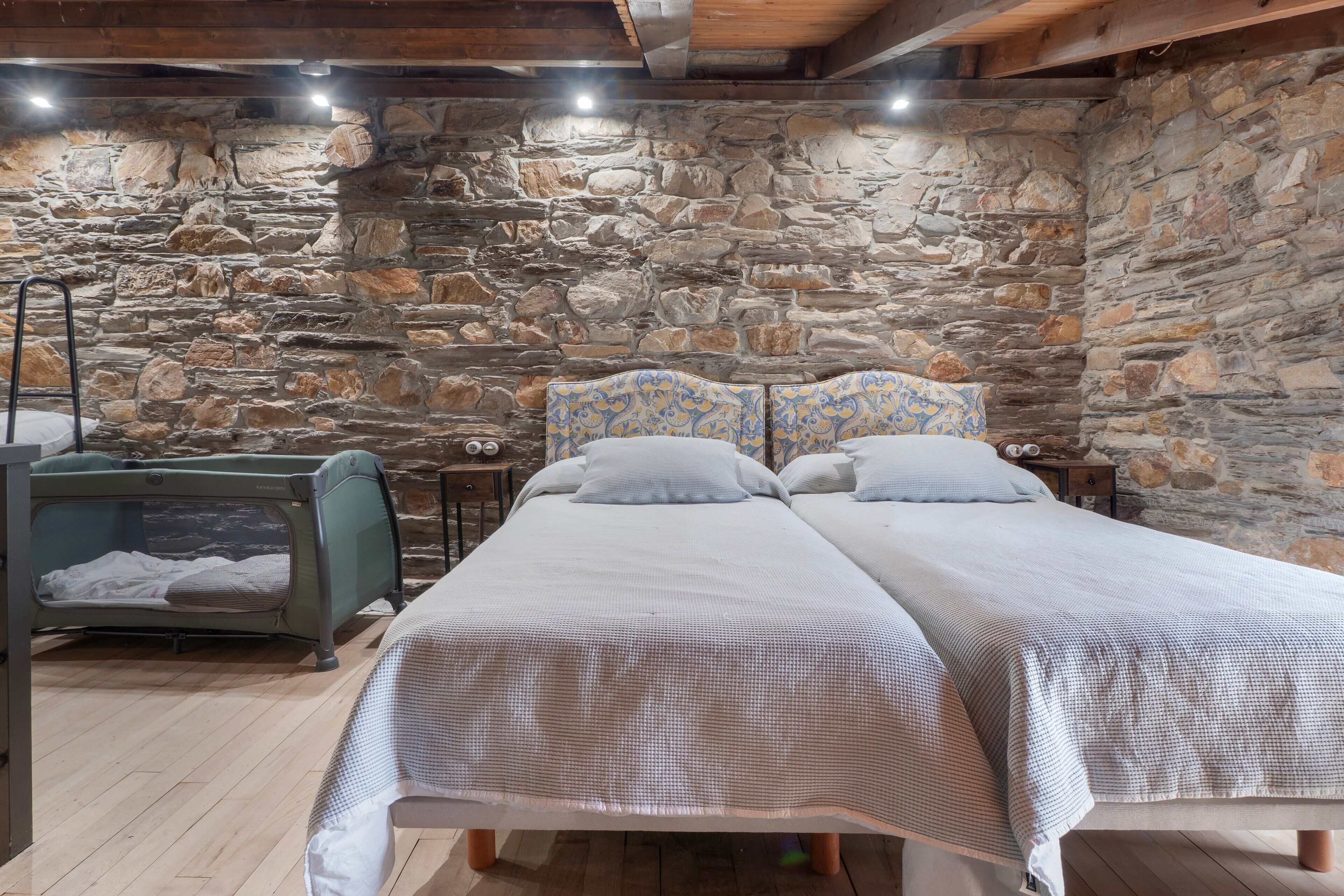 A bedroom with a stone wall, a bed with a patterned headboard, a suitcase, and a small table with a clock.