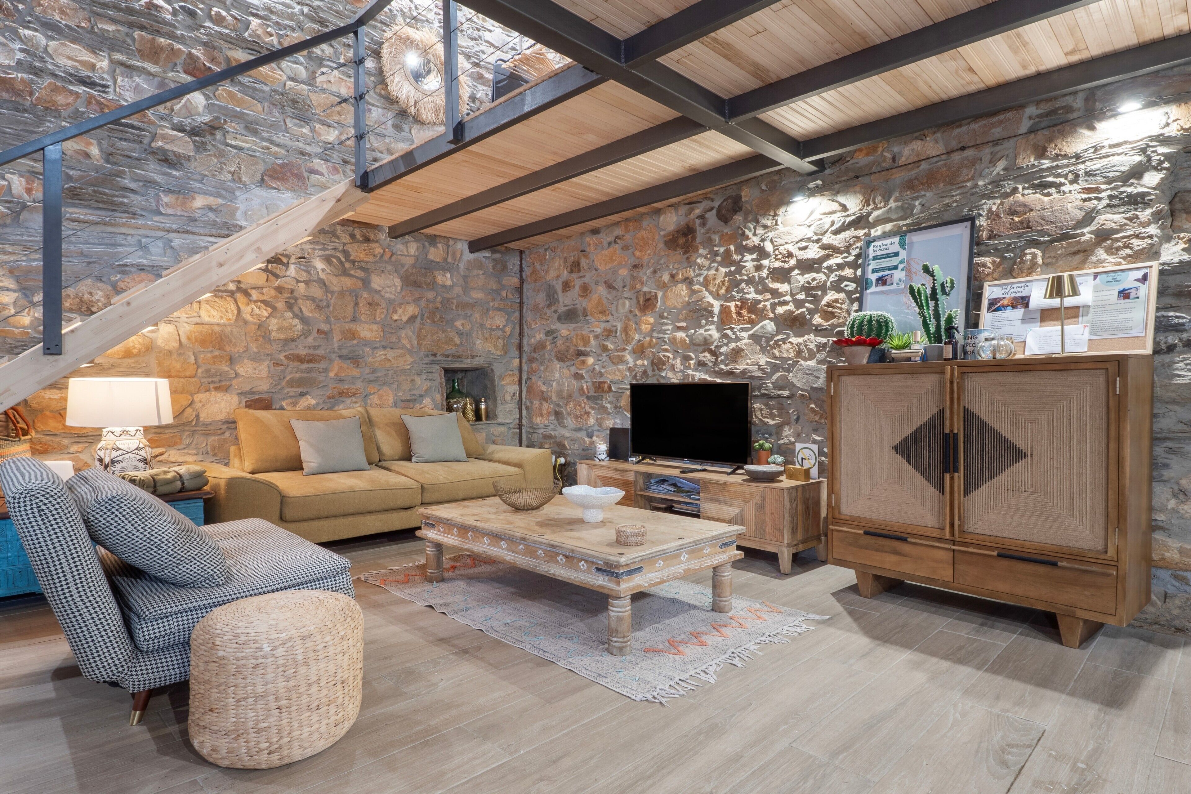 Una sala rústica con una pared de piedra, una escalera de madera, un sofá, un televisor y un aparador.