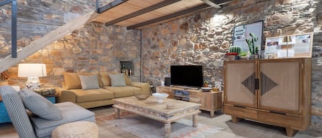 A rustic living room with a stone wall, a wooden staircase, a sofa, a television, and a cabinet.