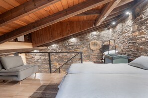 Un dormitorio acogedor en un ático con pared de piedra, vigas de madera, una cama, un sofá y una lámpara colgante.