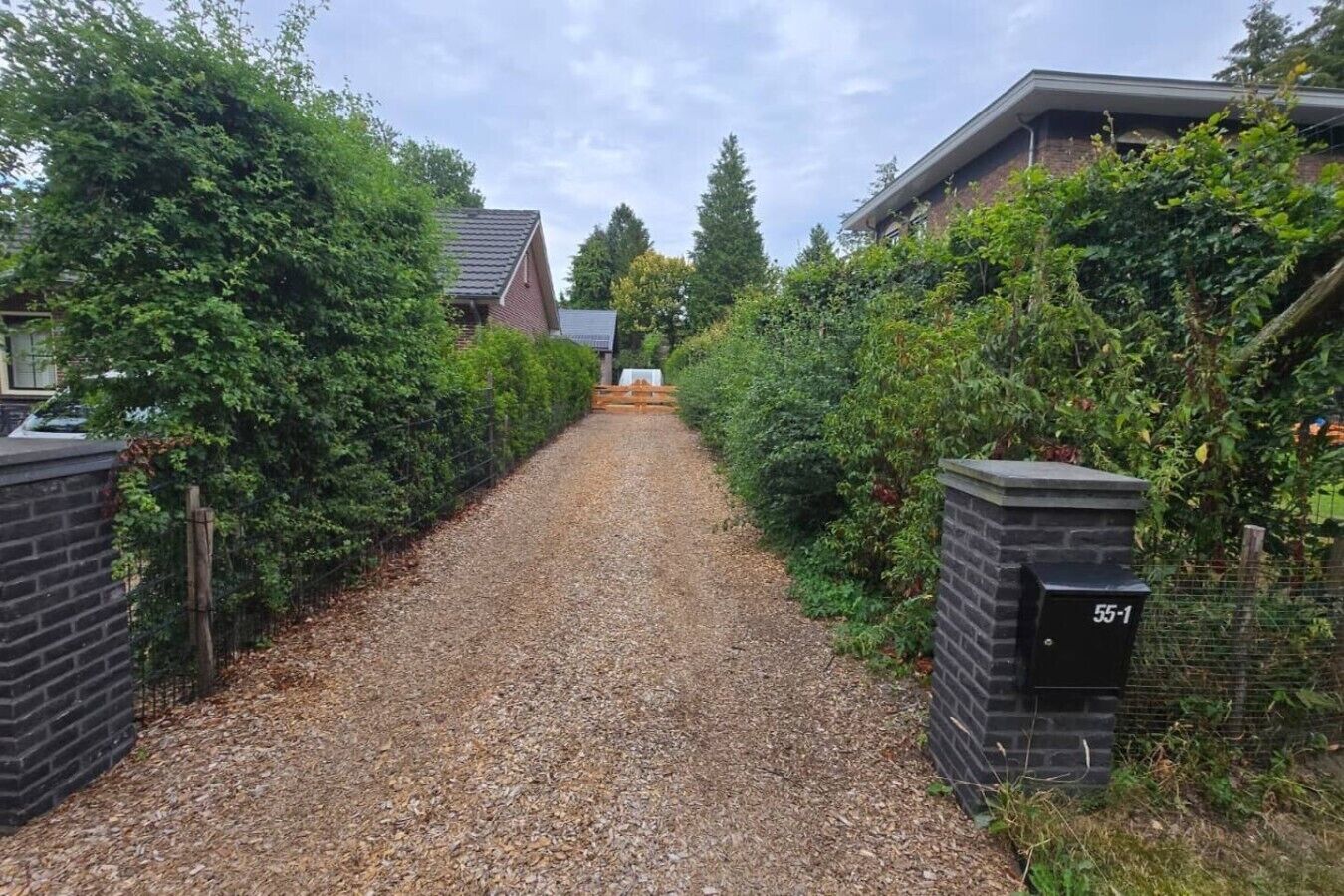En grusuppfart leder fram till ett hus med en mur av tegelsten på ena sidan och en grind av metall på den andra.
