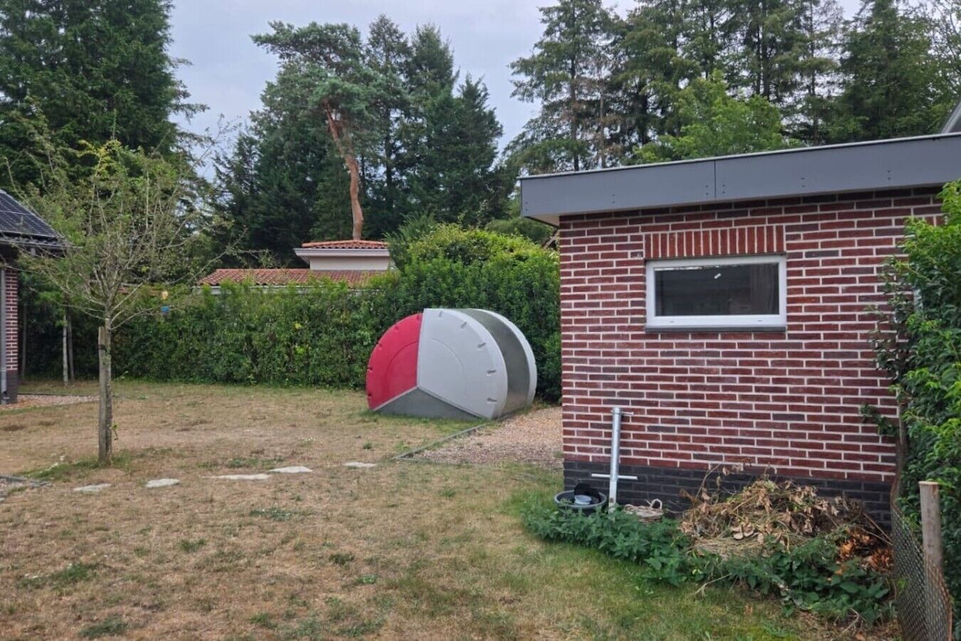 A brick building with a window, a red and white object in the yard, and a tree.