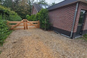 A gravel driveway leading to a wooden gate, a brick building with a window, and a house in the background. - Eendje 1 | Vrijstaande 6-persoons vakantiewoning (Ermelo)