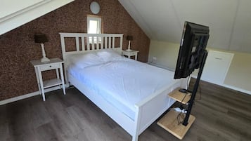 A bedroom with a sloped ceiling, a bed, a television on a stand, and a small bedside table with a lamp.