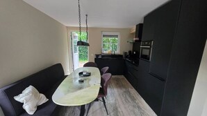 A compact kitchen with a dining area, a black sofa, and a window providing natural light. - Eendje 1 | Vrijstaande 6-persoons vakantiewoning (Ermelo)