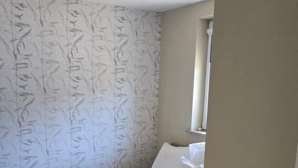 A bedroom with a bed, a patterned wallpaper, a window, and a ceiling light fixture. - Eendje 1 | Vrijstaande 6-persoons vakantiewoning (Ermelo)