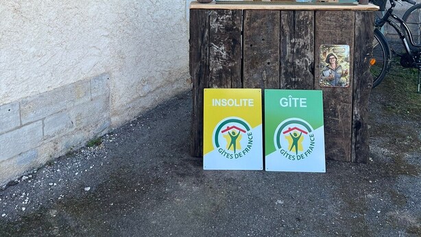 A rustic outdoor setting with a wooden table, a bicycle, and two signs indicating 'INSOLITE' and 'GÎTE' services.