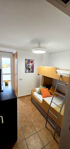 A dormitory room with a bunk bed, a desk, a chair, a ladder, a ceiling fan, and a window with a view.