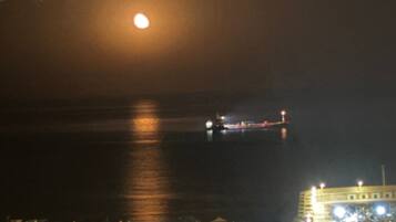 Vista costera nocturna con una luna destacada, un barco distante y edificios iluminados.
