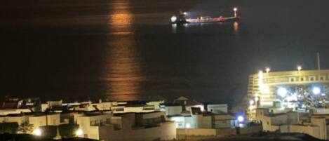 Une vue côtière de nuit, avec une lune bien visible, un navire au loin et des bâtiments illuminés.