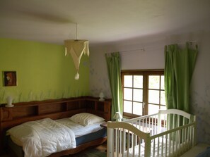 A room with a bed, a crib, a window with curtains, and a hanging light fixture. - Cottage "Le Moulin de Laval" with Private Terrace, Wi-Fi, and Air Conditioning (Saint-Priest-des-Champs)