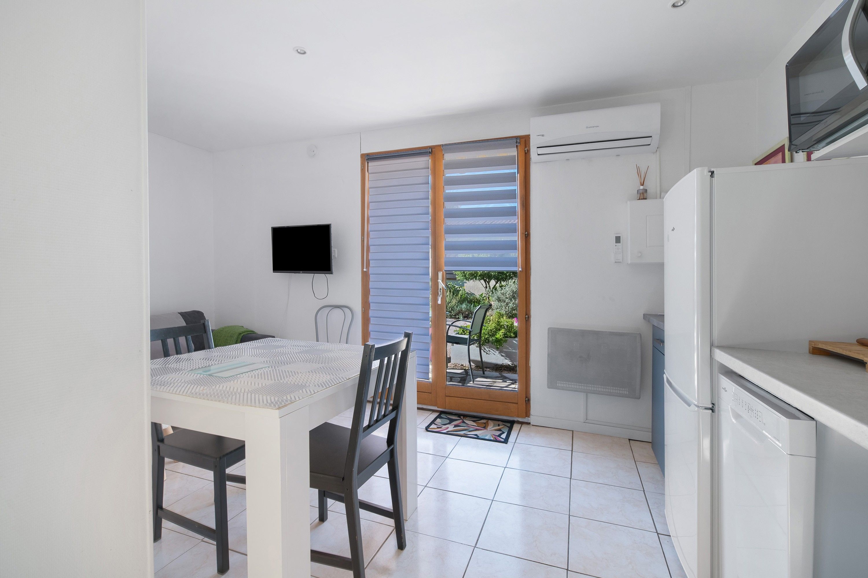 A compact kitchen with a refrigerator, a dining table with chairs, a television mounted on the wall, and a sliding glass door leading to an outdoor area.