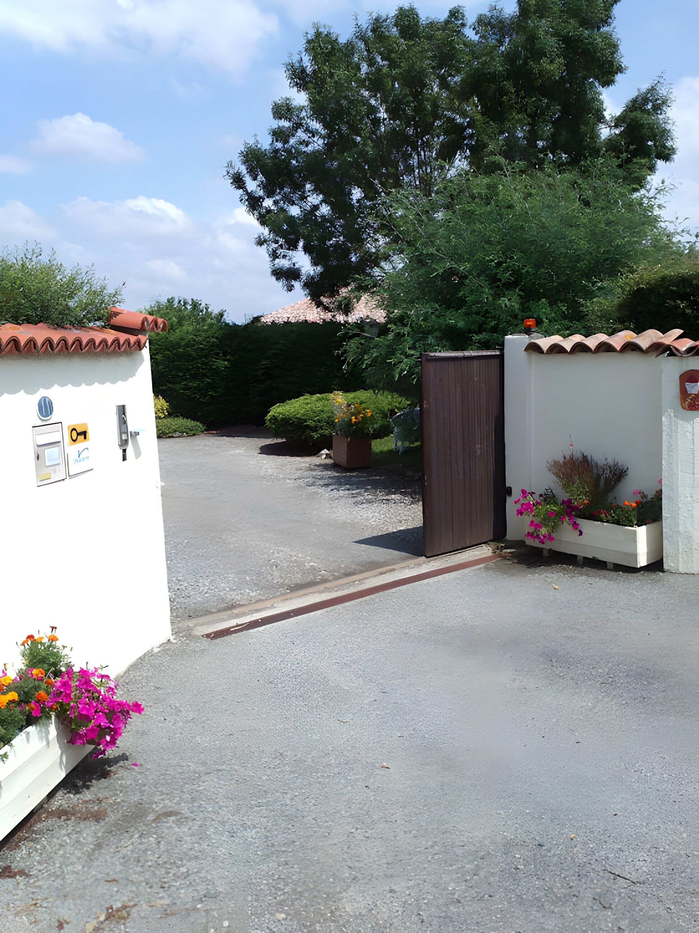 Una entrada con puerta de reja, un edificio blanco, un camino de grava y macetas con flores.