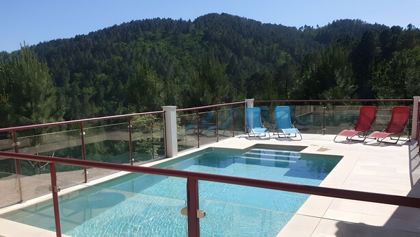 Ein Swimmingpool mit Glasgeländern, roten und blauen Liegestühlen und Blick auf einen bewaldeten Berg.