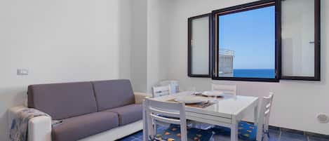 A small, modern room with a sofa, a dining table with chairs, and a large window offering a view of the sea.