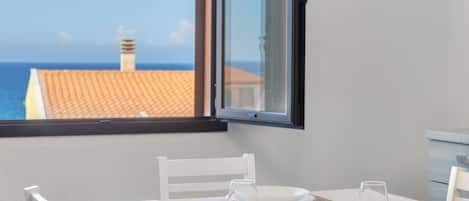 A dining table with chairs, a window with a view of the sea and buildings, and a table setting with plates, glasses, and cutlery.