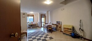 A room with a checkered floor, a dining table with chairs, a small kitchen area, and a vacuum cleaner.