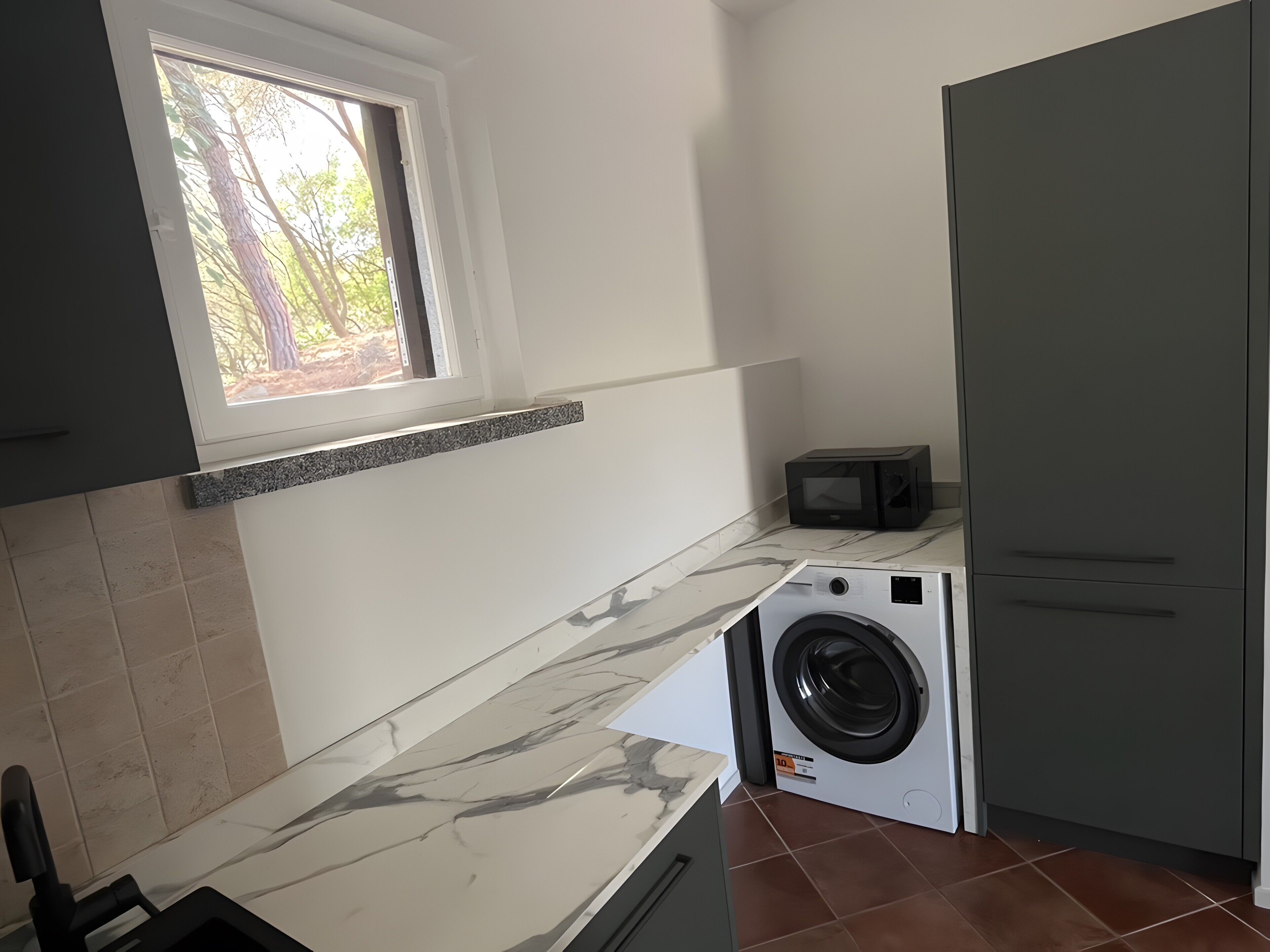Una cucina compatta con lavatrice, microonde e finestra vista alberi.