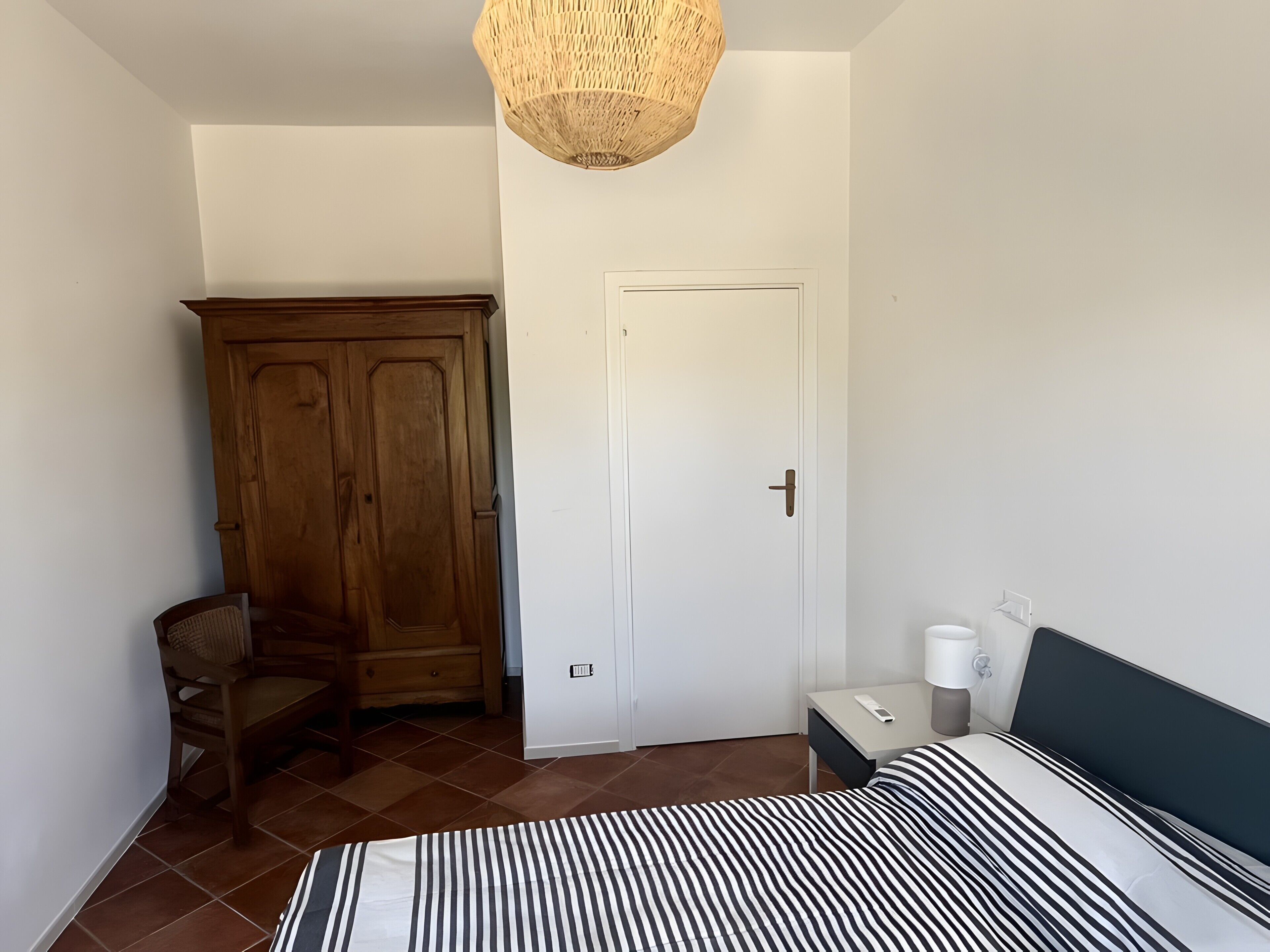 A bedroom with a bed, a wardrobe, a chair, a bedside table with a lamp, and a hanging light fixture.