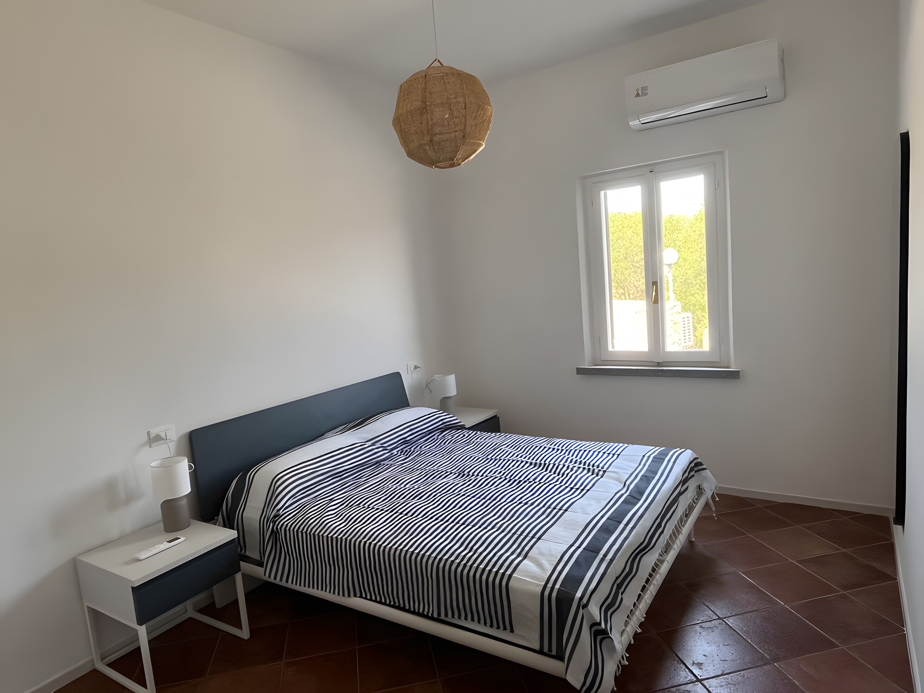 A bedroom with a bed, bedside tables, a hanging light, a window, and an air conditioning unit.
