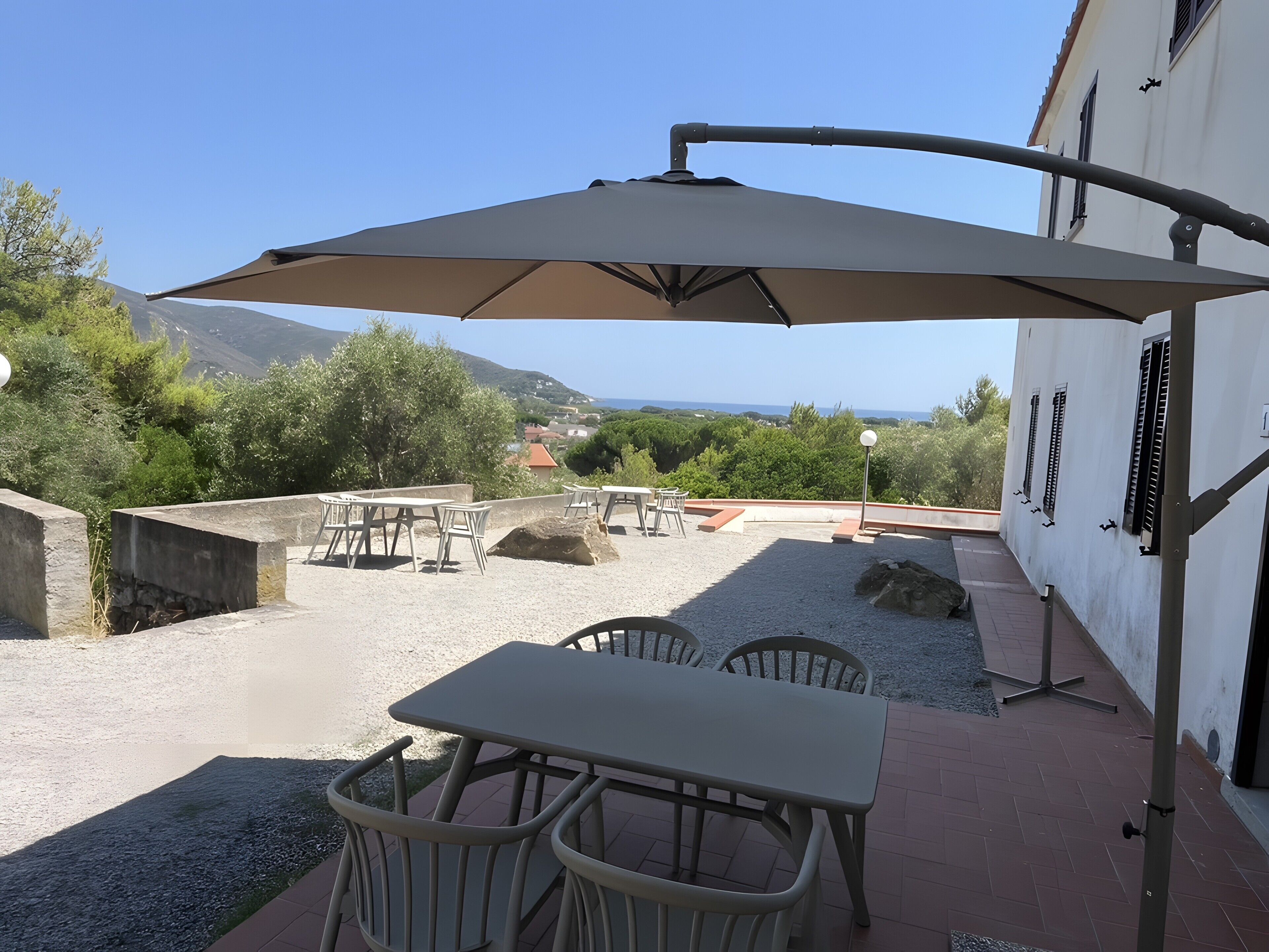 An outdoor patio with tables and chairs, a large umbrella, and a view of the sea and hills.