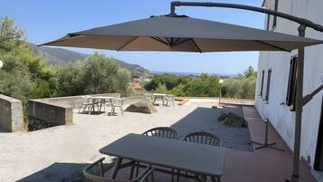 An outdoor patio with tables and chairs, a large umbrella, and a view of the sea and hills.