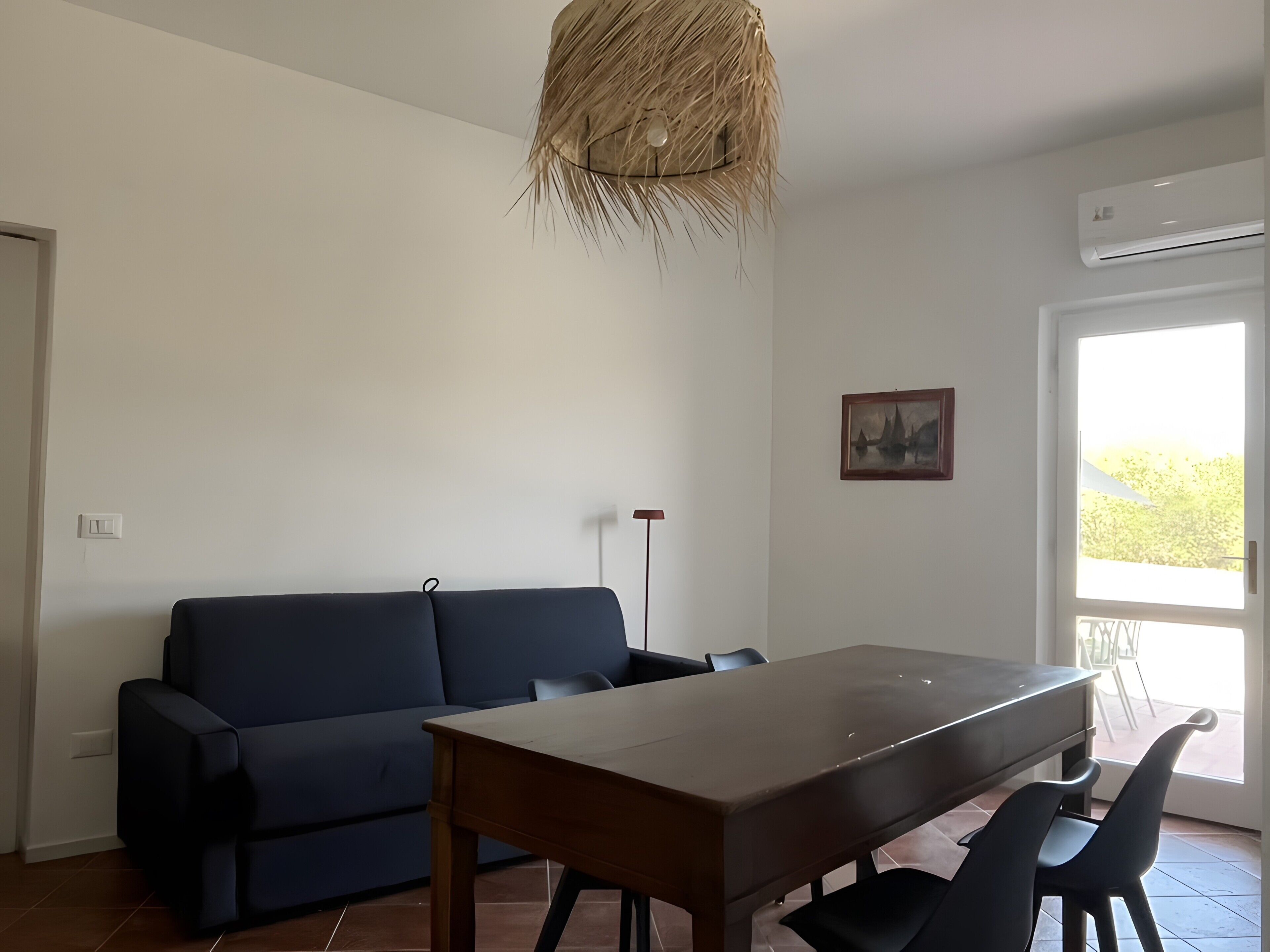 Una habitación con un sofá azul oscuro, una mesa de comedor de madera y dos sillas negras. Hay una lámpara en el techo y un cuadro en la pared.