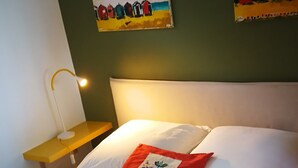 A neatly made bed with white linens, a red throw pillow, and a small table with a lamp beside it.