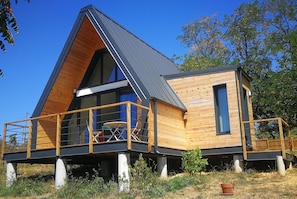 Una cabaña moderna de madera con techo triangular, un balcón con mesa y sillas, y un ventanal pequeño.