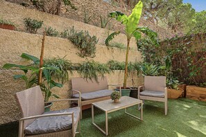 Zona d'estar a l'aire lliure amb mobiliari de vimet i una taula petita, envoltada de diverses plantes en testos i una tanca de bambú.