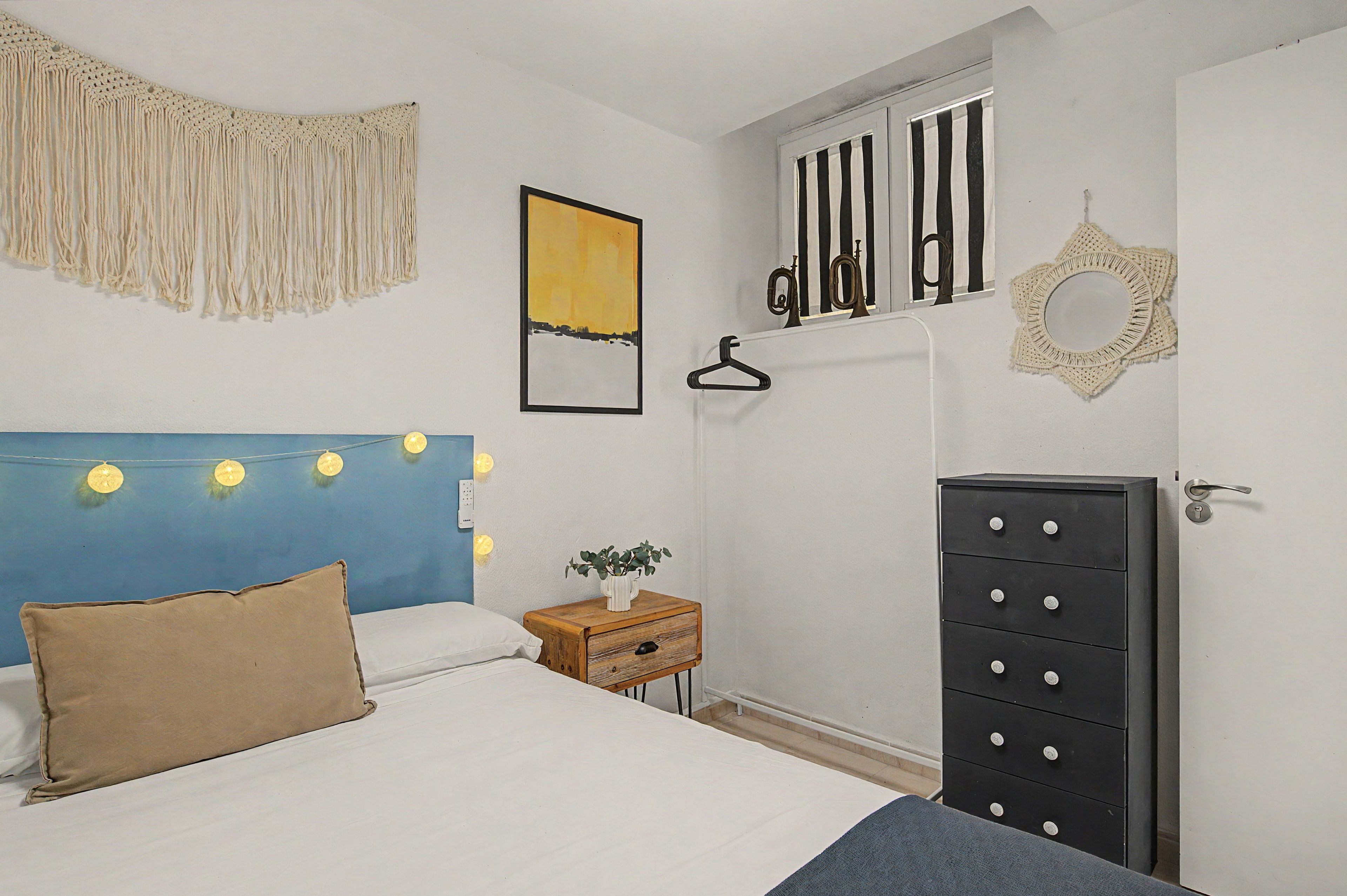 A bedroom with a bed, a dresser, a nightstand, a wall-mounted light fixture, and a hanging macrame piece.