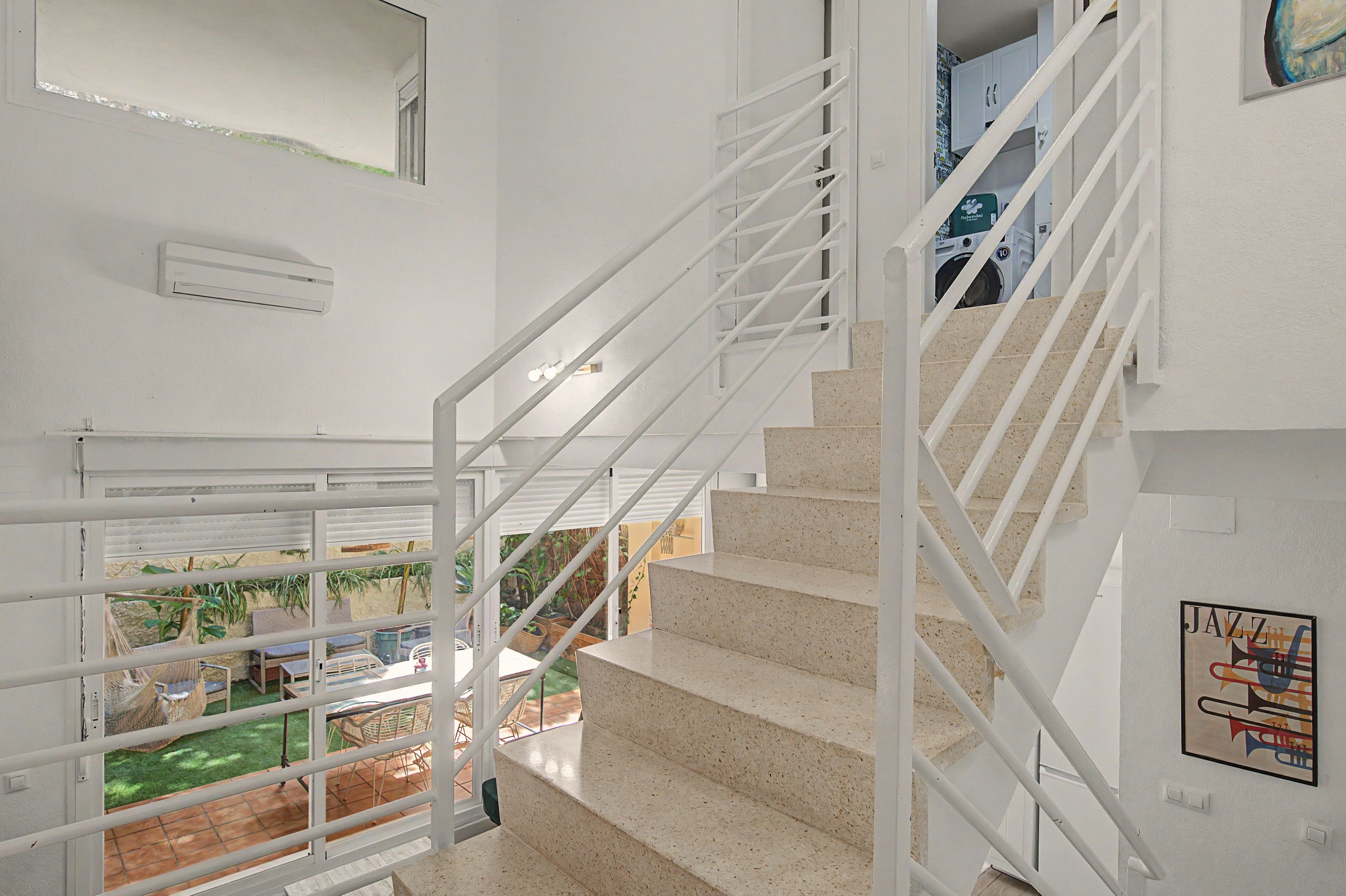 Una escalera con barandillas blancas que conduce a un segundo piso, vista de un jardín exterior y un aire acondicionado mural.