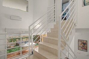 Una escalera con barandillas blancas que conduce a un segundo piso, vista de un jardín exterior y un aire acondicionado mural.