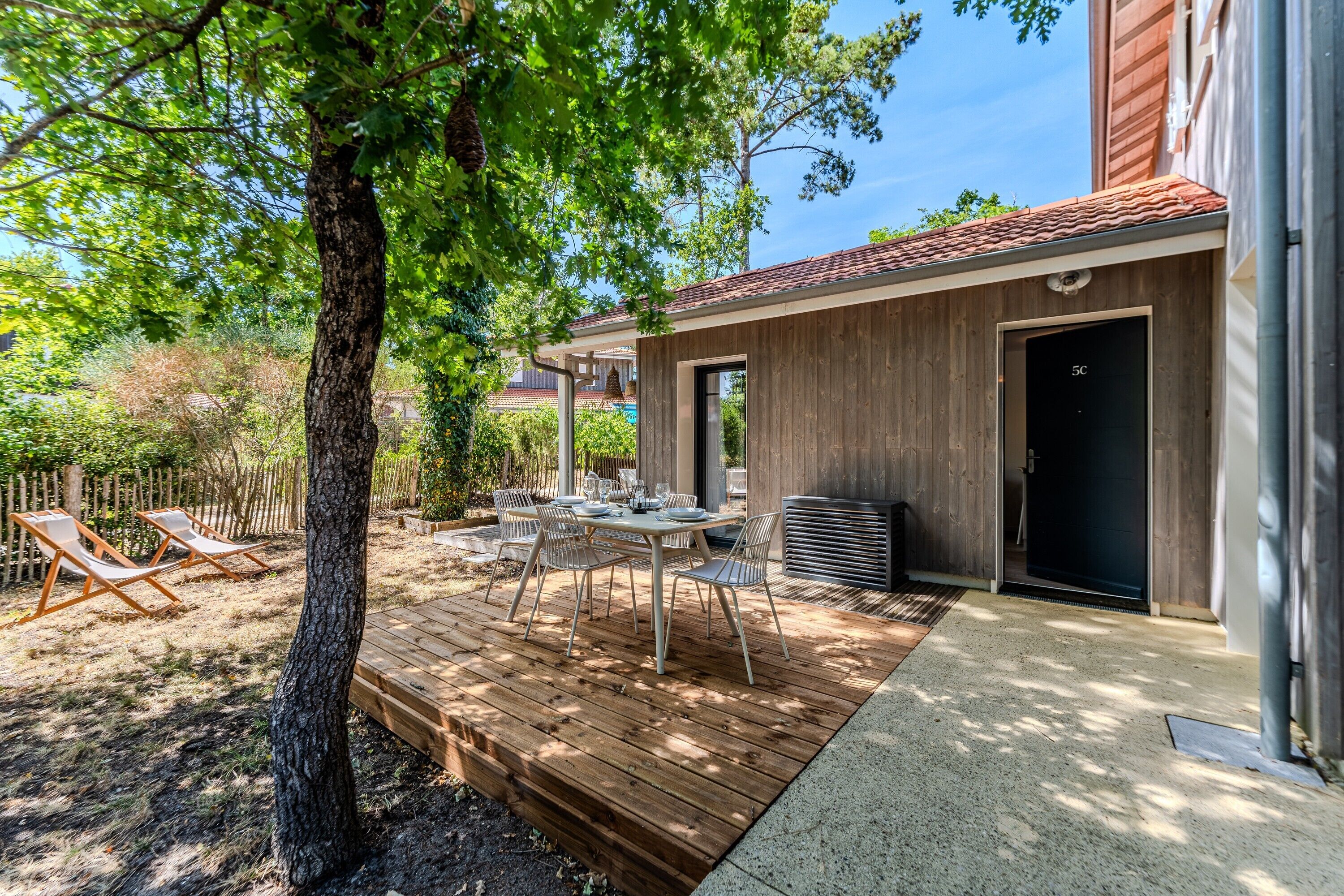 Une terrasse en bois avec une table et des chaises, un espace extérieur couvert et un bâtiment doté d’une porte sombre.