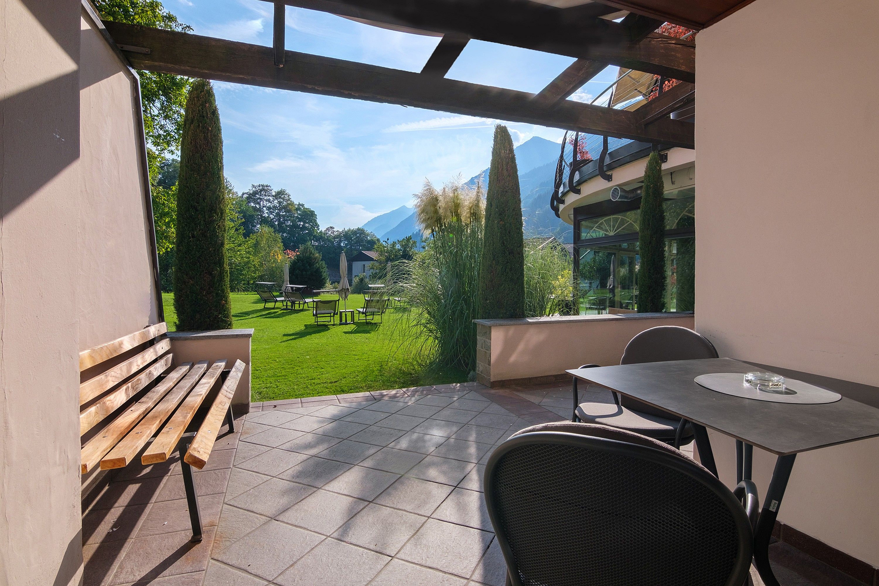A patio with a wooden bench, a table, and chairs, overlooking a well-maintained lawn and distant mountains.
