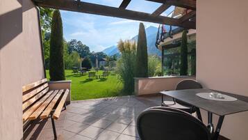 A patio with a wooden bench, a table, and chairs, overlooking a well-maintained lawn and distant mountains.