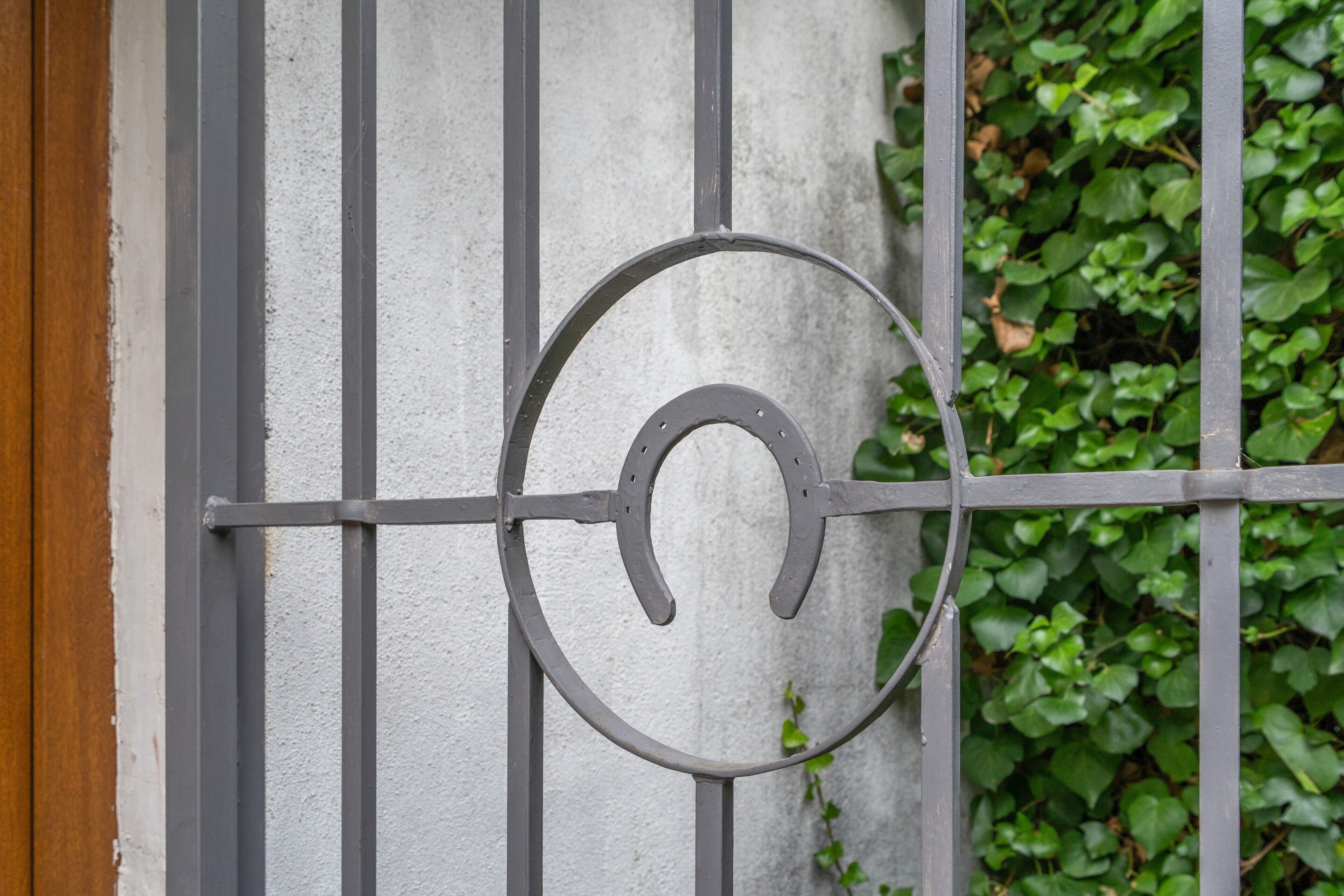 Una reja metálica con un emblema circular, frente a un muro con textura y rodeada de follaje.